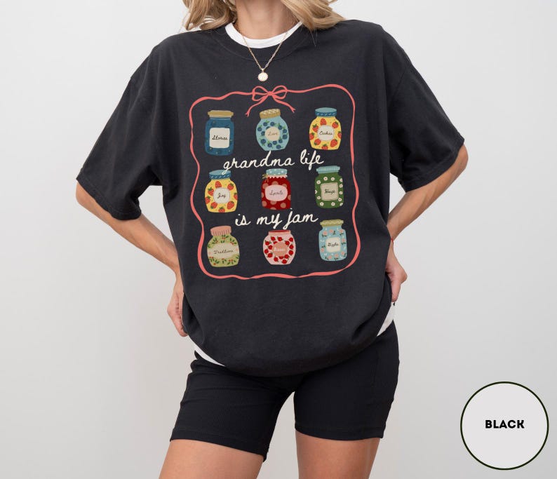 Peut inclure: T-shirt noir avec un motif de neuf pots de confiture dans un cadre rose. Le texte "grandma life is my jam" est &eacute;crit en police script. Le t-shirt est de coupe d&eacute;contract&eacute;e et &agrave; manches courtes.