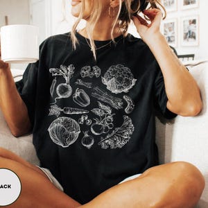 May include: Black t-shirt featuring a detailed white illustration of various vegetables, including carrots, beets, and cauliflower. The design is a botanical drawing style. The shirt is a relaxed fit.
