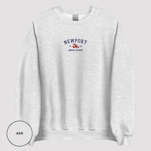 May include: Light grey sweatshirt featuring the word "NEWPORT" in navy blue arched text, a red lobster graphic, and the text "16 39 RHODE ISLAND". The sweatshirt is a Gildan brand.