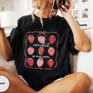 Puede incluir: Camiseta negra con un gr&aacute;fico de fresas y el texto "crochet is my jam". El gr&aacute;fico presenta nueve fresas en varios tonos de rojo y rosa, enmarcadas por un borde rosa con un lazo en la parte superior. La camiseta es de corte holgado.