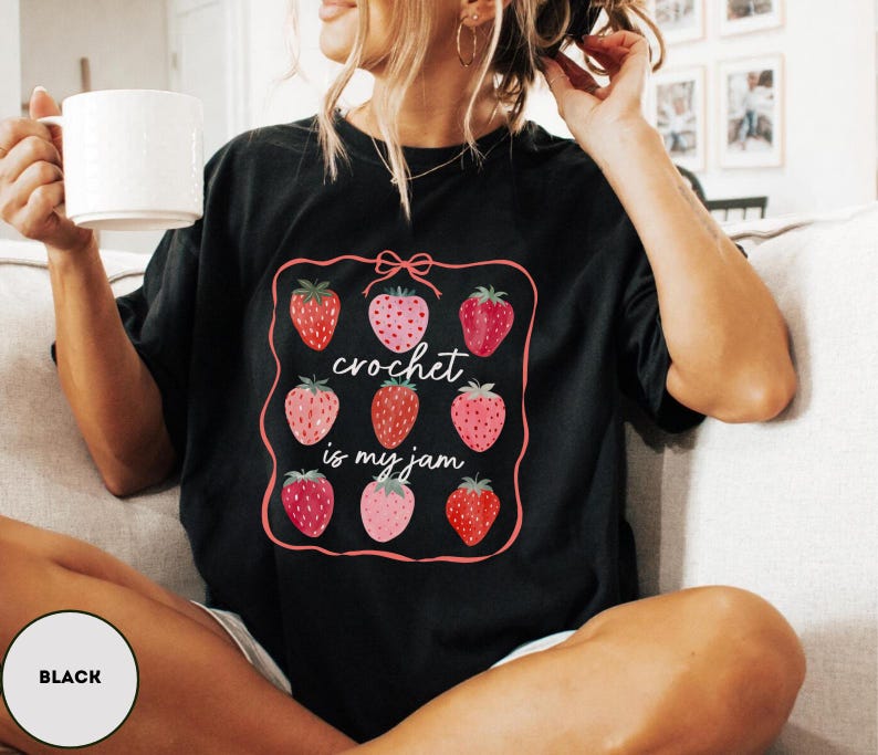 Puede incluir: Camiseta negra con un gr&aacute;fico de fresas y el texto "crochet is my jam". El gr&aacute;fico presenta nueve fresas en varios tonos de rojo y rosa, enmarcadas por un borde rosa con un lazo en la parte superior. La camiseta es de corte holgado.