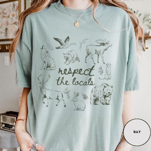 May include: A light blue t-shirt featuring a collection of wildlife illustrations, including a wolf, bear, moose, and rabbit. The phrase "respect the locals" is printed in a handwritten style below the animal graphics.