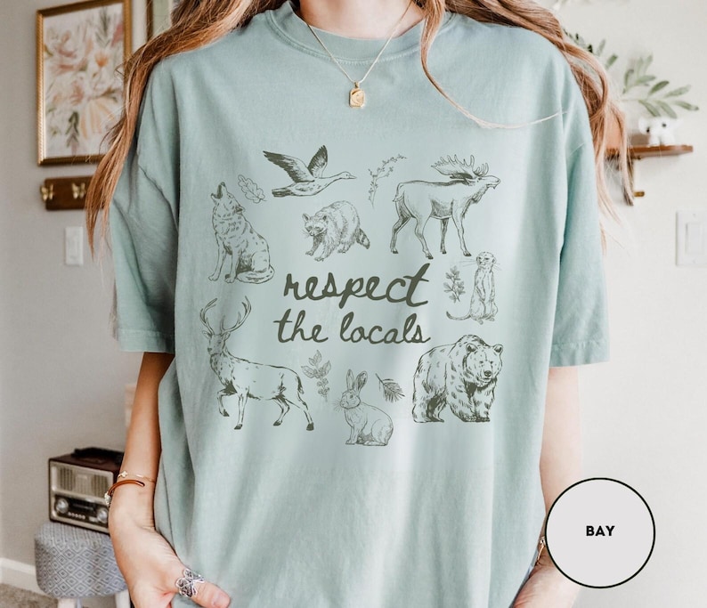 May include: A light blue t-shirt featuring a collection of wildlife illustrations, including a wolf, bear, moose, and rabbit. The phrase "respect the locals" is printed in a handwritten style below the animal graphics.