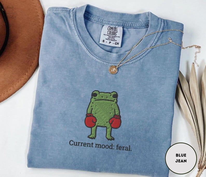 May include: A blue jean colored t-shirt with a green frog wearing red boxing gloves. The text "Current mood: feral" is below the frog. A gold necklace with a pendant is on the shirt.