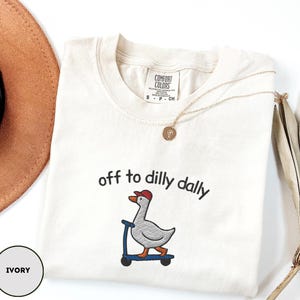May include: Ivory-colored t-shirt with the phrase "off to dilly dally" above an embroidered goose wearing a red cap and riding a blue scooter. A gold necklace and a brown hat are also visible.