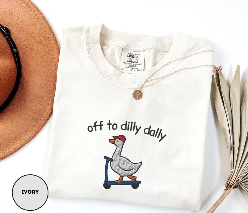 May include: Ivory-colored t-shirt with the phrase "off to dilly dally" above an embroidered goose wearing a red cap and riding a blue scooter. A gold necklace and a brown hat are also visible.