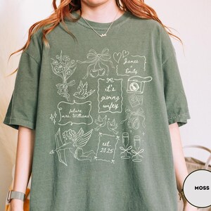 May include: Moss green t-shirt with white graphic illustrations. The design includes floral elements, doves, a camera, champagne glasses, and the text "it's giving wifey". Additional text includes "future mrs. Williams" and "est. 2025".
