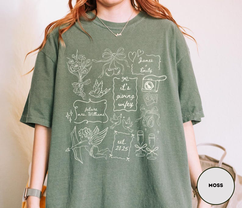 May include: Moss green t-shirt with white graphic illustrations. The design includes floral elements, doves, a camera, champagne glasses, and the text "it's giving wifey". Additional text includes "future mrs. Williams" and "est. 2025".