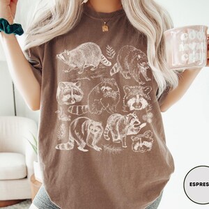 Op de afbeelding: Espresso-kleurig T-shirt met witte wasbeer illustraties. Het ontwerp toont verschillende poses van wasberen en botanische details. Een comfortabel, casual kledingstuk.