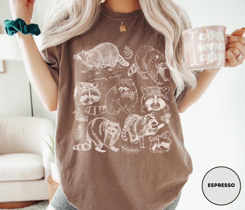 Op de afbeelding: Espresso-kleurig T-shirt met witte wasbeer illustraties. Het ontwerp toont verschillende poses van wasberen en botanische details. Een comfortabel, casual kledingstuk.