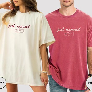May include: Ivory and crimson oversized t-shirts. Each shirt features the text "just meowied" above a graphic of two cats. The ivory shirt is worn by a person, and the crimson shirt is worn by another person. The shirts are made of a soft material.