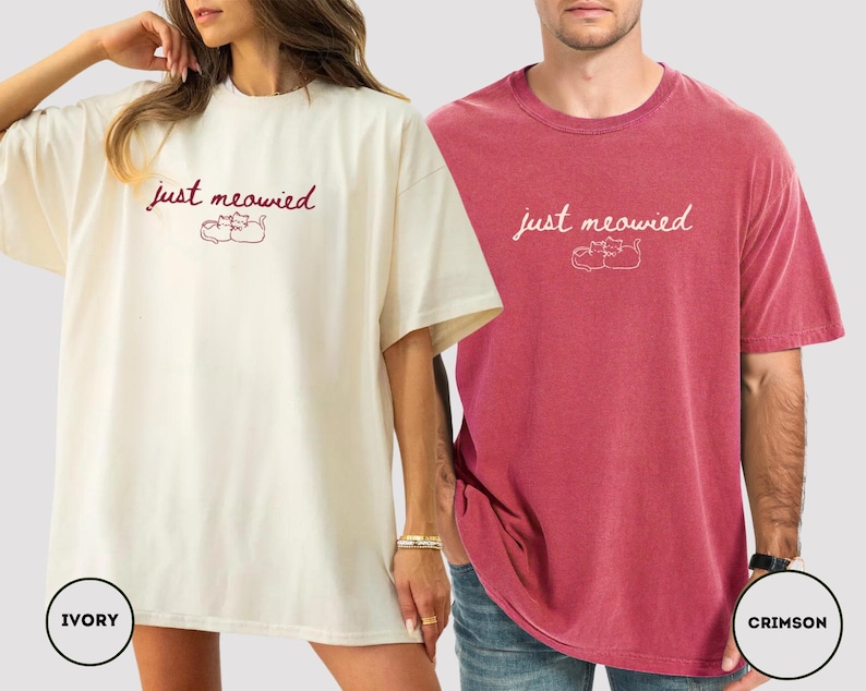 May include: Ivory and crimson oversized t-shirts. Each shirt features the text "just meowied" above a graphic of two cats. The ivory shirt is worn by a person, and the crimson shirt is worn by another person. The shirts are made of a soft material.