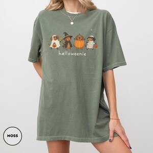 Op de afbeelding: Een mosgroen T-shirt met korte mouwen en een afbeelding van vier teckels in Halloween-kostuums, waaronder een geest, een heks, een pompoen en een mummie, met de tekst "halloweenie" in het wit onder de honden gedrukt.