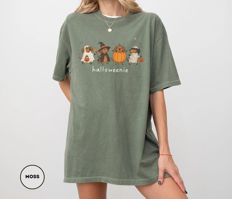 Op de afbeelding: Een mosgroen T-shirt met korte mouwen en een afbeelding van vier teckels in Halloween-kostuums, waaronder een geest, een heks, een pompoen en een mummie, met de tekst "halloweenie" in het wit onder de honden gedrukt.