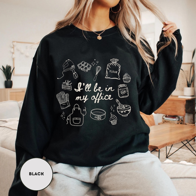 May include: Black sweatshirt with white line art of baking tools and the text "I'll be in my office." The design includes a mixer, whisk, flour sack, and other kitchen items. A round label at the bottom left says "BLACK."