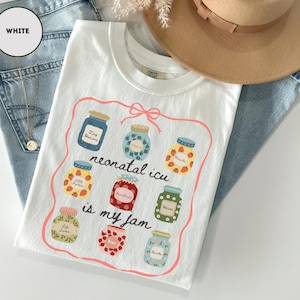 May include: White t-shirt with a colorful graphic of jam jars and the text "neonatal icu is my jam". The shirt is folded on top of blue jeans, with a tan hat and a gold necklace visible in the background.