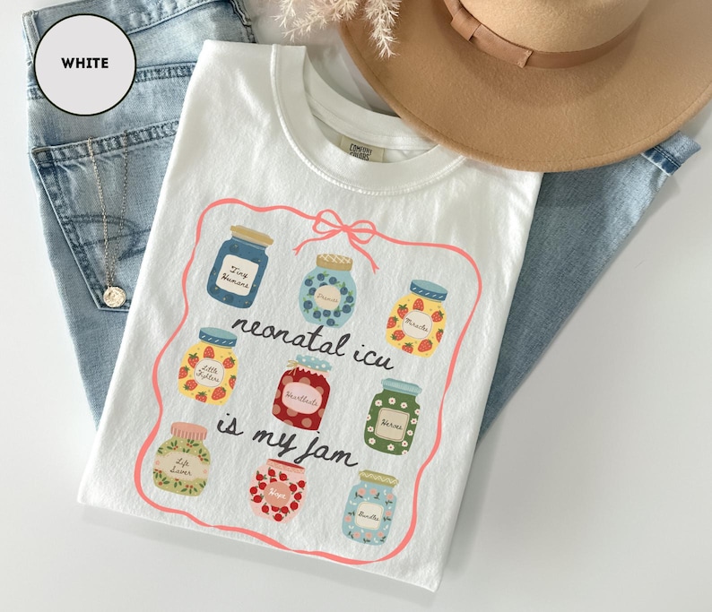 May include: White t-shirt with a colorful graphic of jam jars and the text "neonatal icu is my jam". The shirt is folded on top of blue jeans, with a tan hat and a gold necklace visible in the background.