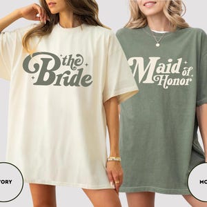 May include: Two oversized t-shirts, one ivory and one moss green. The ivory shirt has "the Bride" in a retro font, and the moss green shirt says "Maid of Honor". Both are short-sleeved and have a relaxed fit, ideal for a bridal party.