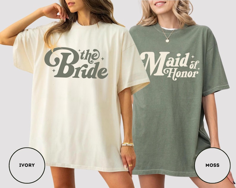 May include: Two oversized t-shirts, one ivory and one moss green. The ivory shirt has "the Bride" in a retro font, and the moss green shirt says "Maid of Honor". Both are short-sleeved and have a relaxed fit, ideal for a bridal party.