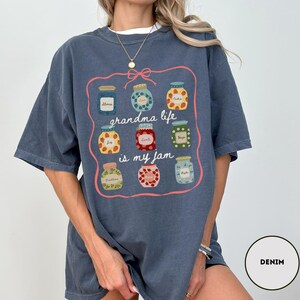 Peut inclure: T-shirt bleu denim avec un motif de bocaux de confiture et le texte "grandma life is my jam". Les bocaux sont remplis de diff&eacute;rents fruits et conserves, soulign&eacute;s d'une bordure rose. Le t-shirt est de coupe ample.