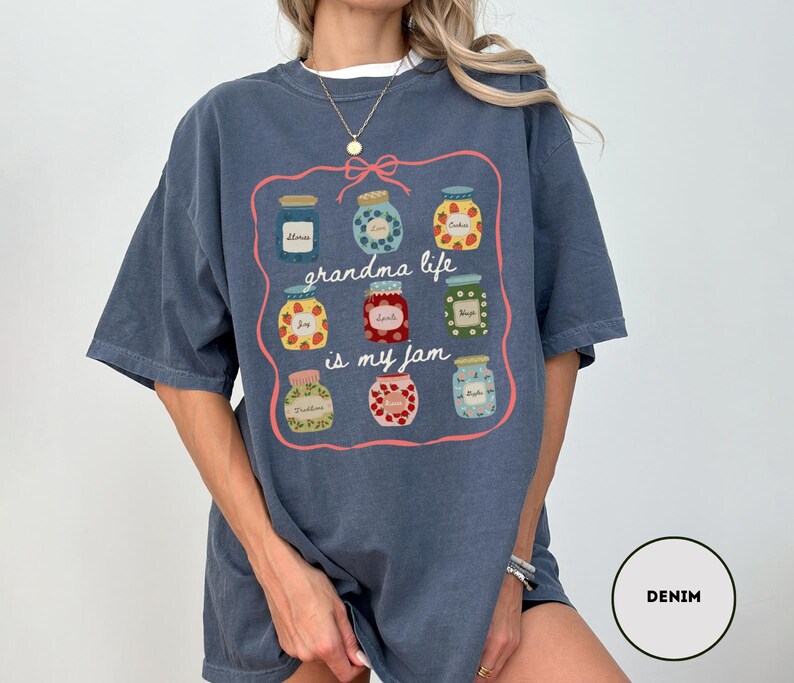 Peut inclure: T-shirt bleu denim avec un motif de bocaux de confiture et le texte "grandma life is my jam". Les bocaux sont remplis de diff&eacute;rents fruits et conserves, soulign&eacute;s d'une bordure rose. Le t-shirt est de coupe ample.