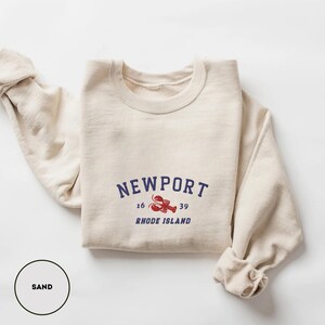 May include: A sand-coloured sweatshirt featuring the word "NEWPORT" in navy blue, with a red lobster graphic and the words "RHODE ISLAND" below. The sweatshirt has rolled-up sleeves.