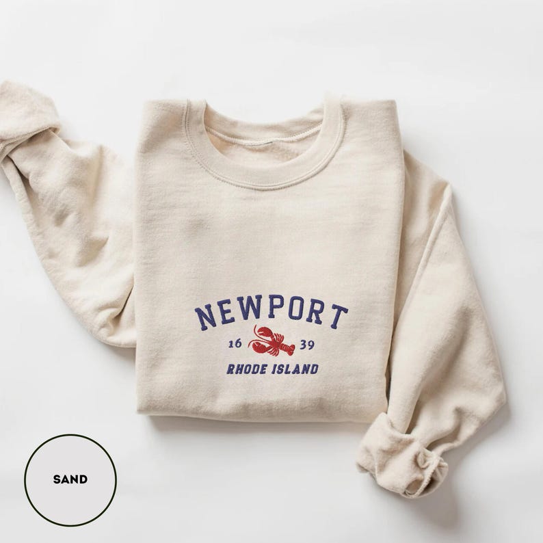 May include: A sand-coloured sweatshirt featuring the word "NEWPORT" in navy blue, with a red lobster graphic and the words "RHODE ISLAND" below. The sweatshirt has rolled-up sleeves.