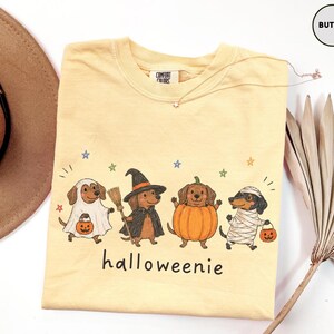 Op de afbeelding: Een botergeel t-shirt met een afbeelding van vier teckels in Halloween-kostuums: een spook, een heks met een bezem, een pompoen en een mummie. Elke hond houdt een pompoenemmer vast. De tekst "halloweenie" staat onder de honden gedrukt.
