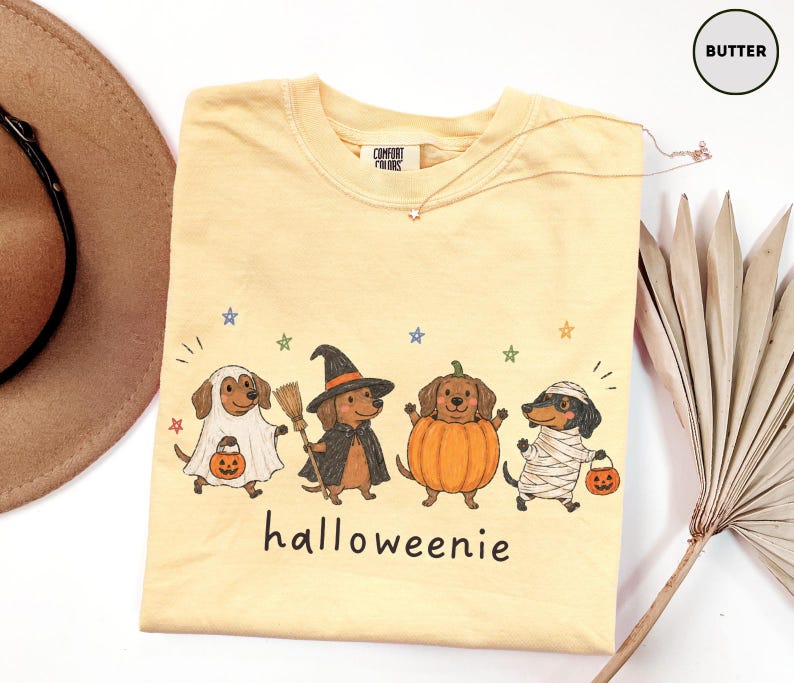 Op de afbeelding: Een botergeel t-shirt met een afbeelding van vier teckels in Halloween-kostuums: een spook, een heks met een bezem, een pompoen en een mummie. Elke hond houdt een pompoenemmer vast. De tekst "halloweenie" staat onder de honden gedrukt.