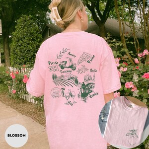 May include: Pink t-shirt with a green graphic of Italian imagery, including a scooter, pizza, and the words "Pasta," "Tuscany," "Rome," and "Venice." The shirt also features a small lemon graphic on the front. The word "Blossom" is at the bottom.