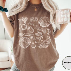 May include: Espresso-colored t-shirt featuring a detailed white illustration of various vegetables, including a beet, carrot, and cauliflower. The shirt has a relaxed fit and is paired with denim shorts. A mug with the text "Cool Mom Club" is held in the hand.