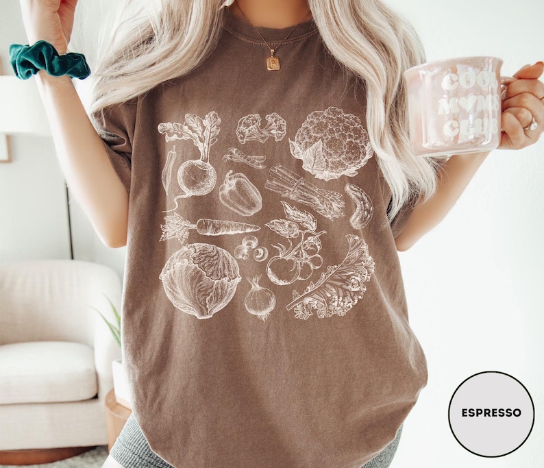 May include: Espresso-colored t-shirt featuring a detailed white illustration of various vegetables, including a beet, carrot, and cauliflower. The shirt has a relaxed fit and is paired with denim shorts. A mug with the text "Cool Mom Club" is held in the hand.