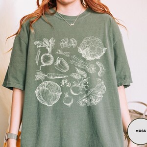 May include: Moss green t-shirt featuring a detailed white illustration of various vegetables, including a radish, carrot, and cauliflower. The shirt has short sleeves and a relaxed fit. The word "MOSS" is printed on a white circle.