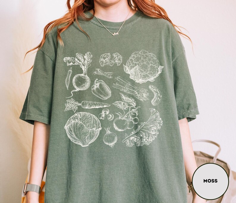 May include: Moss green t-shirt featuring a detailed white illustration of various vegetables, including a radish, carrot, and cauliflower. The shirt has short sleeves and a relaxed fit. The word "MOSS" is printed on a white circle.