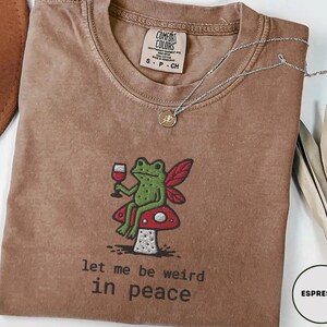 May include: Espresso-colored t-shirt with an embroidered design of a green frog holding a wine glass, seated on a red and white mushroom. The text "let me be weird in peace" is below the frog. A silver necklace is draped across the shirt.