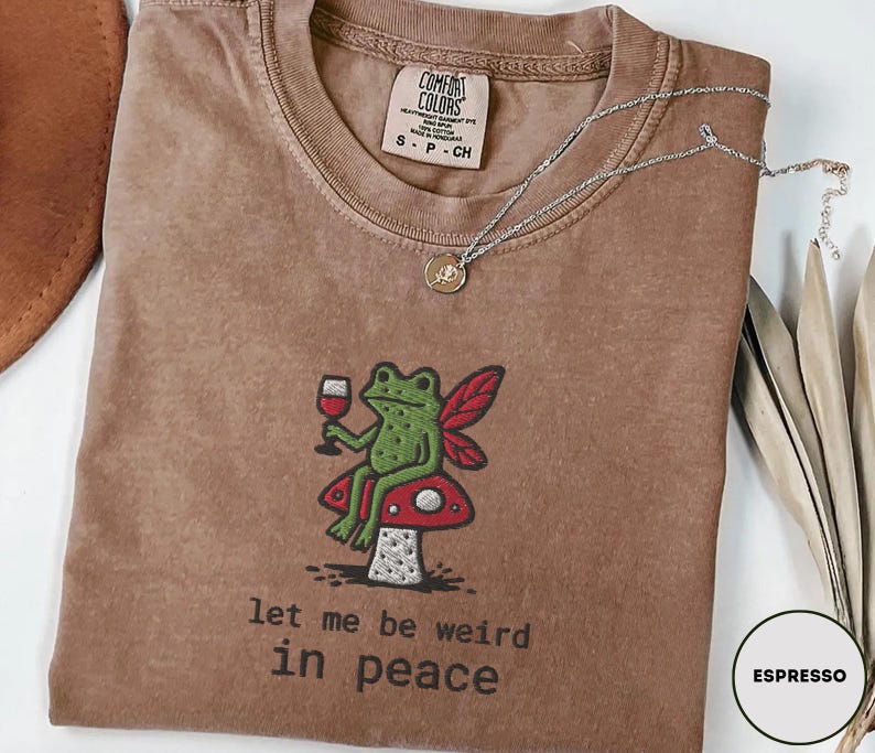 May include: Espresso-colored t-shirt with an embroidered design of a green frog holding a wine glass, seated on a red and white mushroom. The text "let me be weird in peace" is below the frog. A silver necklace is draped across the shirt.