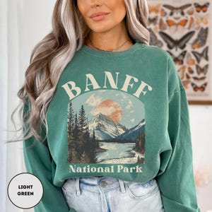 May include: A light green sweatshirt with a vintage-style graphic of a mountain scene and the text "Banff National Park".