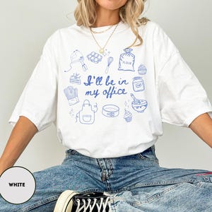 May include: White t-shirt with blue illustrations of baking tools and the phrase "I'll be in my office." The design includes a mixer, whisk, oven mitt, apron, and other baking-related images. The shirt is a relaxed fit.