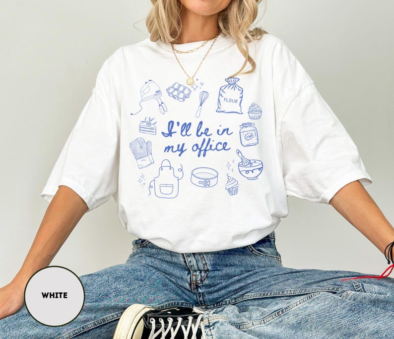 May include: White t-shirt with blue illustrations of baking tools and the phrase "I'll be in my office." The design includes a mixer, whisk, oven mitt, apron, and other baking-related images. The shirt is a relaxed fit.