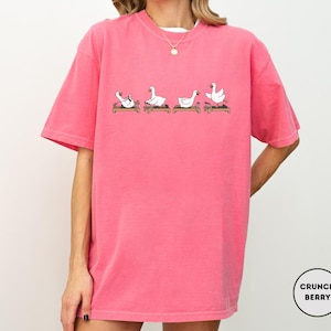 Puede incluir: Camiseta rosa brillante con un dise&ntilde;o de dibujos animados de cuatro gansos blancos en varias posturas de yoga sobre esterillas marrones. La camiseta tiene mangas cortas y cuello redondo. La marca "Crunch Berry" est&aacute; en un c&iacute;rculo en la parte inferior derecha.