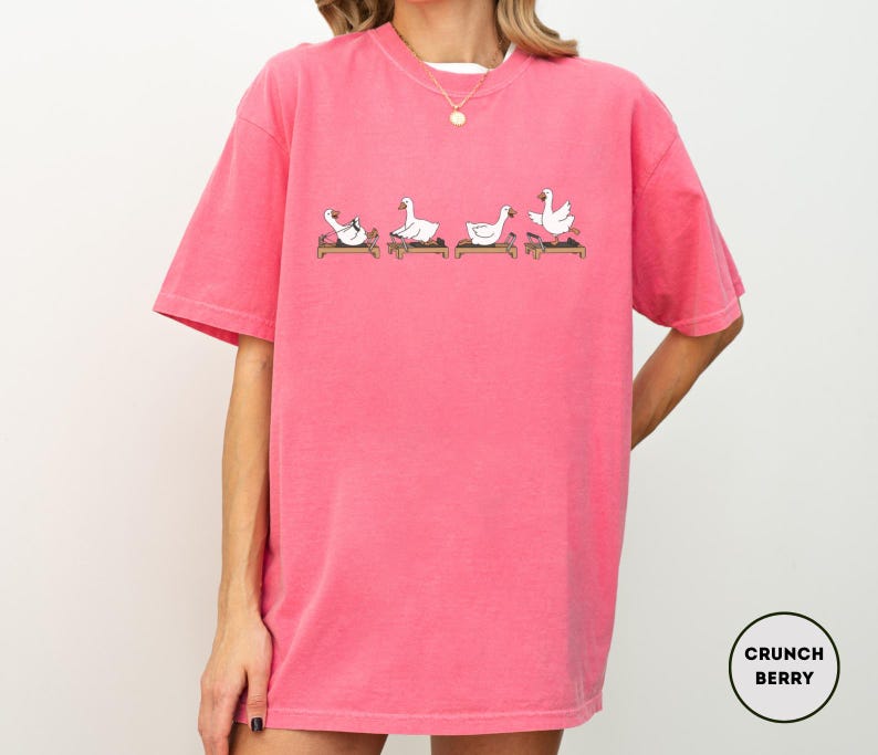 Puede incluir: Camiseta rosa brillante con un dise&ntilde;o de dibujos animados de cuatro gansos blancos en varias posturas de yoga sobre esterillas marrones. La camiseta tiene mangas cortas y cuello redondo. La marca "Crunch Berry" est&aacute; en un c&iacute;rculo en la parte inferior derecha.
