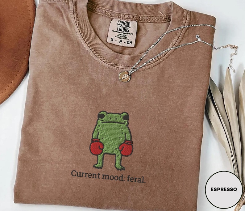 May include: Espresso-colored t-shirt with a green frog wearing red boxing gloves. The text "Current mood: feral" is printed below the frog. A silver necklace with a small pendant is draped across the shirt.
