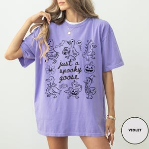 May include: Violet-colored t-shirt with a black graphic of Halloween-themed ducks and the text "just a spooky goose". The design includes a bat, a skull, a pumpkin, and other spooky elements.