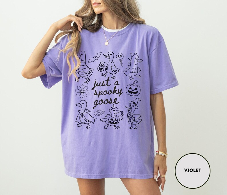 May include: Violet-colored t-shirt with a black graphic of Halloween-themed ducks and the text "just a spooky goose". The design includes a bat, a skull, a pumpkin, and other spooky elements.