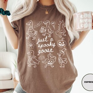 May include: Espresso-colored t-shirt with a white graphic of cartoon geese in Halloween costumes and the text "just a spooky goose". The design includes a bat, pumpkin, and other spooky elements.