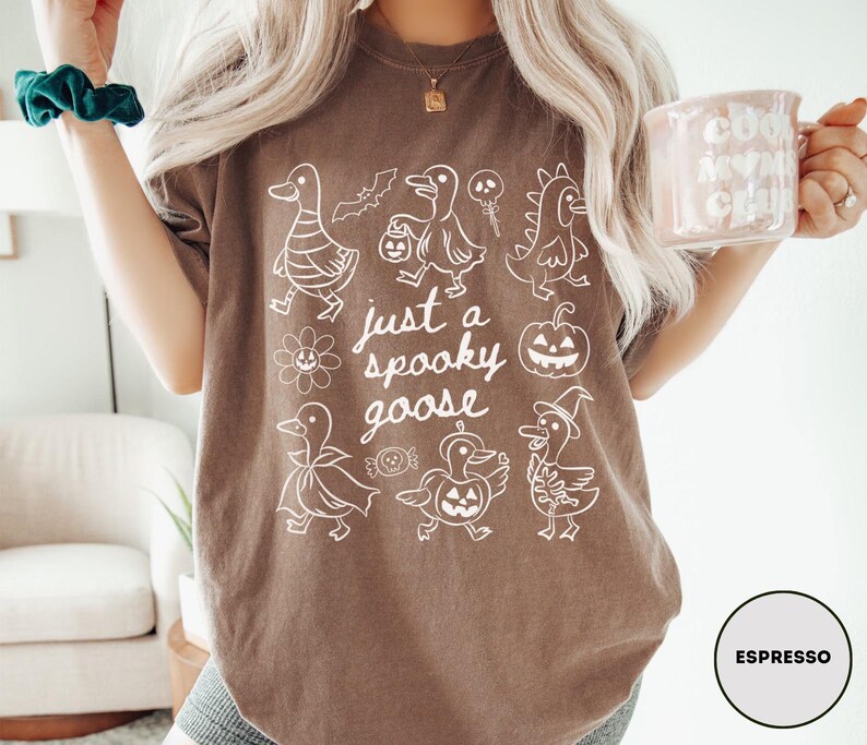 May include: Espresso-colored t-shirt with a white graphic of cartoon geese in Halloween costumes and the text "just a spooky goose". The design includes a bat, pumpkin, and other spooky elements.