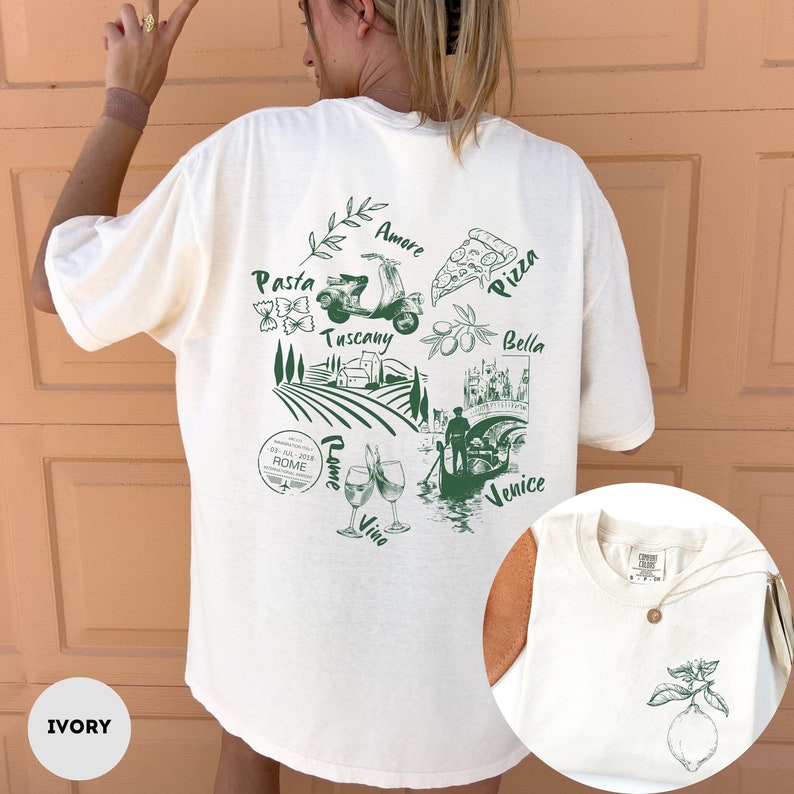 May include: Ivory-colored t-shirt with a green graphic featuring Italian imagery: pasta, pizza, a Vespa, and landmarks. Text includes "Pasta," "Amore," "Pizza," "Tuscany," "Bella," "Rome," "Vino," and "Venice." A small lemon graphic is on the front.