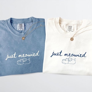 May include: Two folded t-shirts, one blue and one cream, with the phrase "just meowied" and a cat illustration. Each shirt has a gold necklace with a round pendant. The shirts are from Comfort Colors.