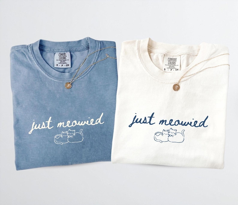 May include: Two folded t-shirts, one blue and one cream, with the phrase "just meowied" and a cat illustration. Each shirt has a gold necklace with a round pendant. The shirts are from Comfort Colors.