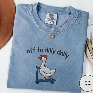 May include: A blue jean colored t-shirt with the phrase "off to dilly dally" embroidered above a cartoon goose riding a scooter. The goose wears a red hat. A gold necklace with a pendant is draped across the shirt.